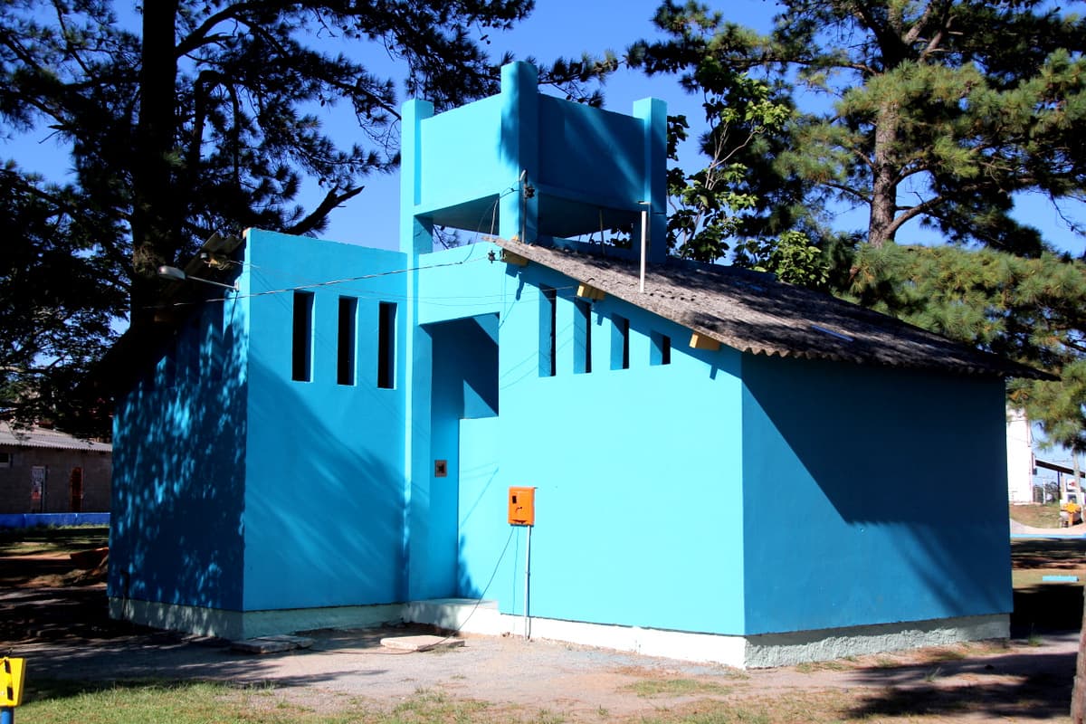 Edificação azul-celeste da Praça Raíssa Vargas, entre pinheiros, em dia de sol